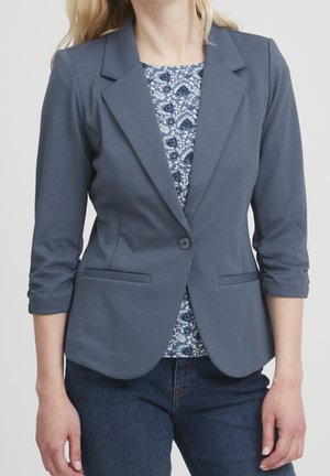 Femme portant un blazer bleu ardoise ajusté avec des manches trois-quarts froncées, par-dessus un top à motif floral bleu et blanc et un jean foncé.