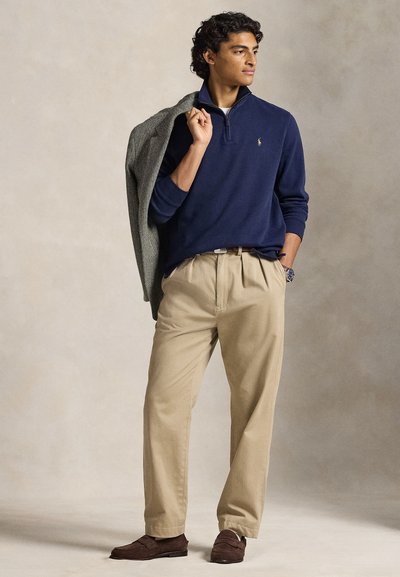 Fiatal férfi tengerészkék negyed cipzáras pulóverben, bézs nadrágban és barna cipőben, egy szürke dzsekit tartva a vállán, egyszínű háttér előtt.