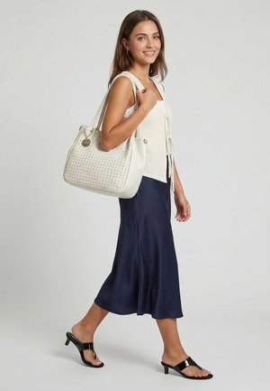 Nuori nainen kävelee kantaen suurta valkoista kuviollista käsilaukkua, päällään kerma liivi, valkoinen toppi, tummansininen hame ja mustat korolliset sandaalit.