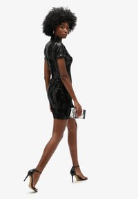 Robe mini noire à paillettes avec des manches courtes, design ajusté et col haut. Associée à des talons hauts noirs avec des lanières et à une pochette métallique.