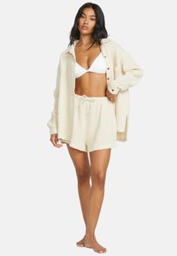 Camicia beige, testurizzata, a maniche lunghe, indossata sopra un bikini bianco e shorts coordinati. Presenta una vita con cordoncino e una chiusura frontale con bottoni.