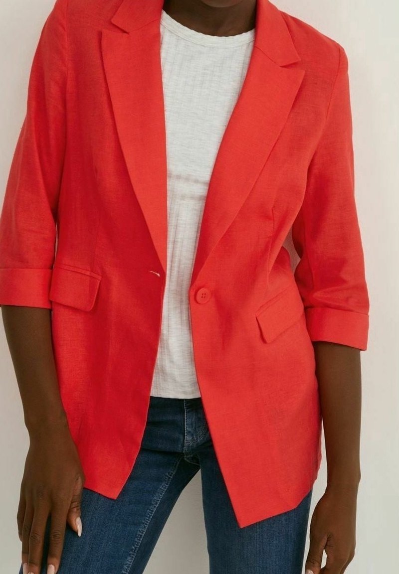 Femme portant un blazer rouge vif avec les manches retroussées, un haut blanc et un jean bleu, debout devant un fond uni.