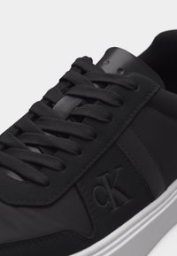 Μαύρο αθλητικό παπούτσι με μαύρα κορδόνια, λείες και υφασμάτινες υφές, ανάγλυφο λογότυπο «ck» στη πλευρά και λευκή λαστιχένια σόλα.