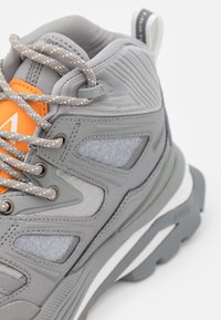 Baskets montantes grises avec une tige texturée, des accents orange et des lacets tressés. Semelle épaisse avec une fine bande blanche et talon renforcé.