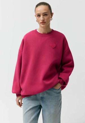 Kobieta z włosami spiętymi do tyłu, nosząca złote kolczyki koła, jasnoróżową, dużych rozmiarów bluzę z detalem w kształcie serca oraz jasnoniebieskie dżinsy, z jedną ręką w kieszeni.