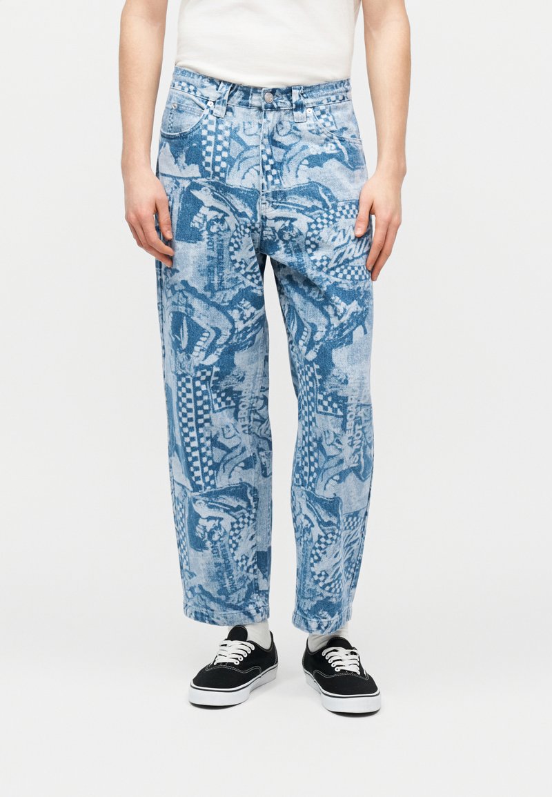 Μπλε τζιν με φαρδιά μπατζάκια και σχέδια, φορεμένα με ασπρόμαυρα αθλητικά παπούτσια και άσπρο τοπ, εμφανίζονται από τη μέση έως τα πόδια.