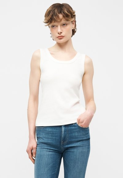 Jeune femme aux cheveux courts portant un haut blanc sans manches et un jean bleu, debout avec une main dans la poche devant un fond uni.