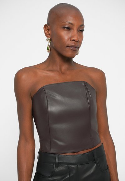 Bruine leren strapless top met een aansluitend ontwerp en een gladde textuur, gecombineerd met grote met groen geaccentueerde oorbellen en zwarte high-waisted broek.