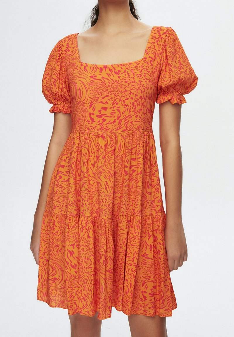 Femme portant une robe à motifs orange et rouges avec des manches courtes bouffantes et un décolleté carré, se tenant devant un fond uni.