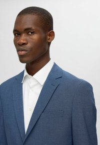 Blazer bleu texturé avec un col cranté, une poche poitrine simple, porté sur une chemise blanche à boutons, affichant une coupe propre et tailleur.