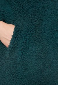 Teal fleecestof met een textuur. Een hand reikt in een zijvak, waardoor het zachte, fluffy materiaal en het comfortabele ontwerp opvallen.