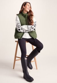 Puffy green vest over white sweater with black patterns, black leggings, and black rubber boots with a logo, seated on a wooden stool.
