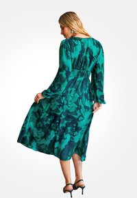 Zielona sukienka w wzór z długimi rękawami, marszczonym talią i zwiewnym dołem. Tkanina ma miękką fakturę z połączeniem ciemnych i jasnych odcieni zieleni.