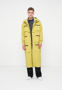 Parka amarela longa com múltiplos bolsos frontais, detalhes com fecho éclair, capuz ajustável e um corte descontraído, usada sobre uma polo cinza e calças pretas.