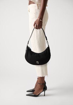 Borsa in ecopelle nera con una forma curva e chiusura con zip, impreziosita da un logo argentato. Il modello indossa jeans chiari a vita alta e tacchi neri.