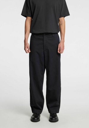 Person iført løse sorte bukser, sort t-shirt og sorte sko, stående mod en ensartet lys baggrund.