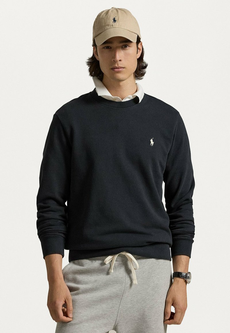 Felpa blu navy con polsini e orlo a coste, con un piccolo logo bianco. Abbinata a pantaloni della tuta grigi e un berretto beige.