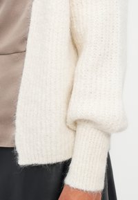 Biały sweter dzianinowy o prążkowanej teksturze, z luźnymi rękawami. Miękki, puszysty materiał, zestawiony z gładką bluzką w kolorze taupe pod spodem.