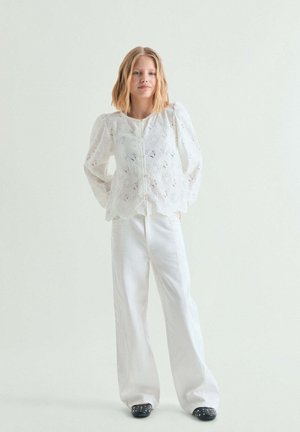 Mujer joven con cabello rubio hasta los hombros, vestida con una blusa blanca de ojales, pantalones blancos de pierna ancha y zapatos negros con tachuelas, de pie contra un fondo liso.