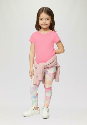 Różowa koszula z krótkim rękawem, jasnoróżowy sweter związany w talii, legginsy w kolorowe serca oraz białe tenisówki.