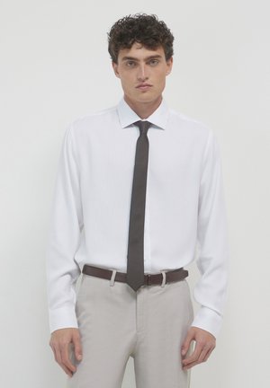 Homme avec des cheveux bouclés portant une chemise blanche, une cravate texturée de couleur foncée, un pantalon beige et une ceinture marron, se tenant devant un fond uni.
