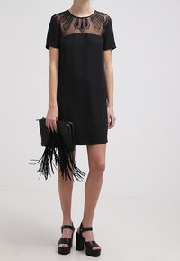 Černé šaty s krátkým rukávem a průhledným vzorovaným výstřihem. Doplnky zahrnují černou kabelku s třásněmi a sandály na silném podpatku.