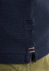 Dámská námořnická modrá košile s waffle texturou, která má tkanou nášivku a světle růžové švy na lemu, v kombinaci s olivově zelenými kalhotami.