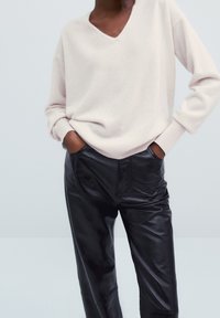 Eine Person trägt einen hellbeigen V-Ausschnitt-Pullover und schwarze Lederhosen, mit den Händen in den Taschen vor einem schlichten hellen Hintergrund.