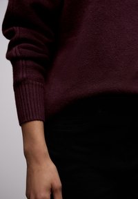Maglione color bordeaux con una finitura morbida e testurizzata, caratterizzato da polsini a costine e una vestibilità rilassata, abbinato a jeans neri in una vista ravvicinata.