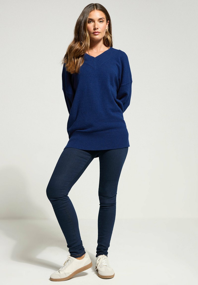 Navyblauer, übergroßer Strickpullover mit V-Ausschnitt, kombiniert mit dunklen Skinny Jeans und weißen Sneakers mit hellbraunen Akzenten.