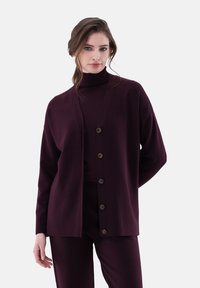 Burgunderfarbener Cardigan aus weichem Material mit V-Ausschnitt, langen Ärmeln und sechs braunen Knöpfen vorne, kombiniert mit passenden Hosen.
