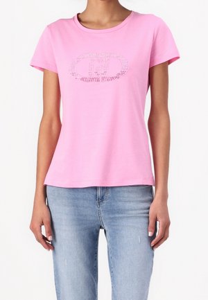 Mujer con camiseta rosa de manga corta con un logo adornado con strass en el pecho y vaqueros azul claro, de pie frente a un fondo blanco.