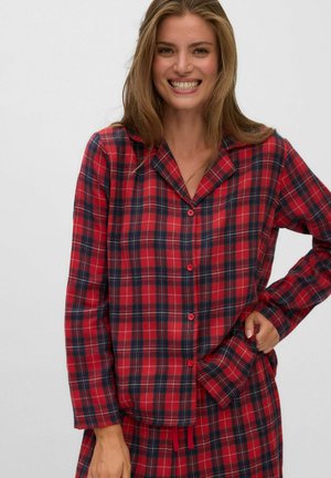 Jeune femme souriante, portant une chemise à manches longues à carreaux rouges et bleu foncé avec boutons et pantalon assorti, debout avec une main sur la hanche.
