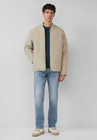 Beżowa pikowana kurtka z okrągłym kołnierzem, długimi r�ękawami i zamkiem z przodu, noszona na ciemnym swetrze i jasnoniebieskich jeansach. Sneakersy z kremowymi akcentami.