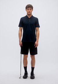 Μαύρο πόλο πουκάμισο με κοντά μανίκια και γιακά, συνδυασμένο με μαύρο σορτς. Το μοντέλο κρατάει ένα γκολφ κλαμπ, φοράει μαύρα παπούτσια και κάλτσες.