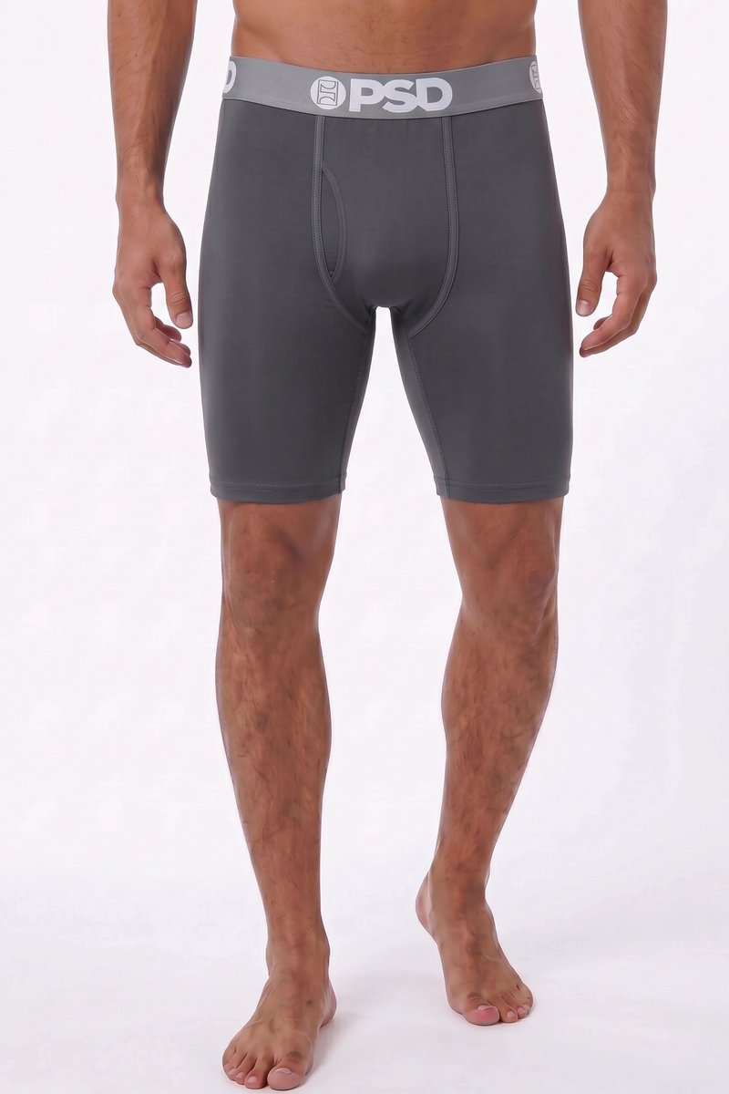 Homme portant un boxer gris foncé de la marque PSD, debout pieds nus sur un fond clair uni avec des jambes et un torse musclés visibles.