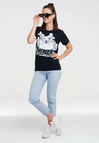 Black cotton T-shirt with Avengers graphic in white, light denim jeans, and white sneakers. Model holds black sunglasses. Plain background.