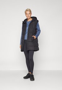 Gilet imbottito nero con cappuccio, caratterizzato da chiusura con zip e due tasche frontali, indossato sopra una maglia a maniche lunghe blu e leggings neri.
