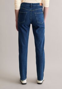 Jean denim bleu foncé à coupe droite, style cinq poches, avec des détails de couture visibles, porté avec des chaussures blanches.