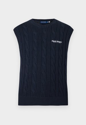 Man met kort blond haar, draagend een wit overhemd, marineblauw kabelgebreid vest, donkere broek, bruine riem, beige loafers, staand met één hand in de zak.