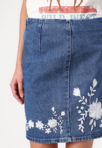Jeans krilo v temno modri barvi z belimi cvetličnimi vezeninami na robu, s pravim krojem in sprednjimi žepi. Klasična tekstura denima.