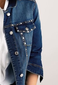 Giubbotto di jeans blu scuro con rivetti e bottoni argento, dotato di una tasca sul petto e maniche arrotolate, abbinato a una camicia bianca sotto.
