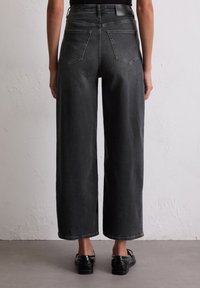 Czarne dżinsy z wysokim stanem i szerokimi nogawkami, z dwoma tylkimi kieszeniami i czystym wykończeniem. Uzupełnione błyszczącymi czarnymi butami.