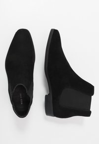 Botas de tornozelo em camurça preta com bico pontiagudo, painéis laterais elásticos e um pequeno salto em borracha. O design é elegante e minimalista.