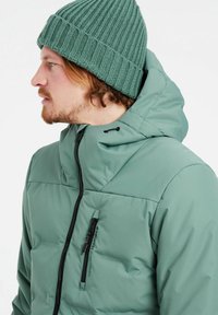 Grüne Puffjacke mit Kapuze, ausgestattet mit einer Reißverschlusstasche und einer glatten, matten Oberfläche. Kombiniert mit einer gerippten grünen Strickmütze.