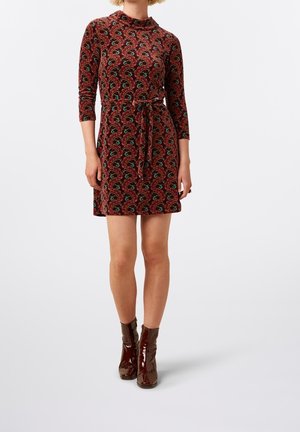Kobieta w czerwonej sukience za kolano z długim rękawem, z kolorowym wzorem w kształcie muszelek i paskiem, w połączeniu z błyszczącymi brązowymi botkami za kostkę.