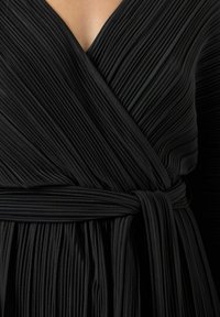 Vestido negro plisado con escote en V profundo, que cuenta con un cinturón atado en la cintura. La tela tiene un aspecto suave y texturizado.