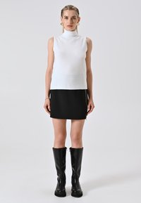Vrouw in een witte mouwloze coltrui, zwarte minirok met een zijrits, zwarte knielange laarzen en gouden oorbellen in een ring, staand tegen een effen achtergrond.