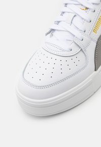 Vit lädersneaker med perforerad tå, grå accenter, platta vita snörningar och en texturerad vit sula med en subtil varumärkeslogotyp i guld.