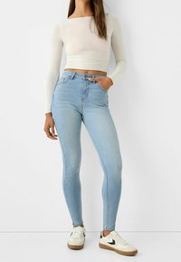 Jasnoniebieskie skinny jeans z wysokim stanem, denimową fakturą, pięcioma kieszeniami i brązowym detalem w postaci guzika; zestawione z dopasowaną białą bluzką z długim rękawem.